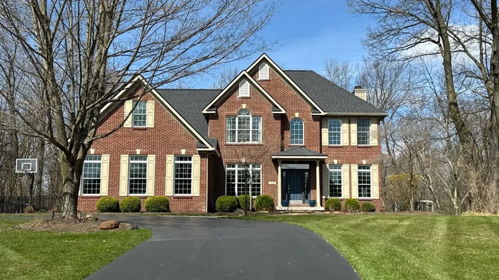 A two-story brick house with yellow shutters, multiple windows, and a paved driveway, surrounded by a front lawn and trees. A basketball hoop is visible on the left side of the driveway. Expert roofing services have ensured the roof's durability and style.