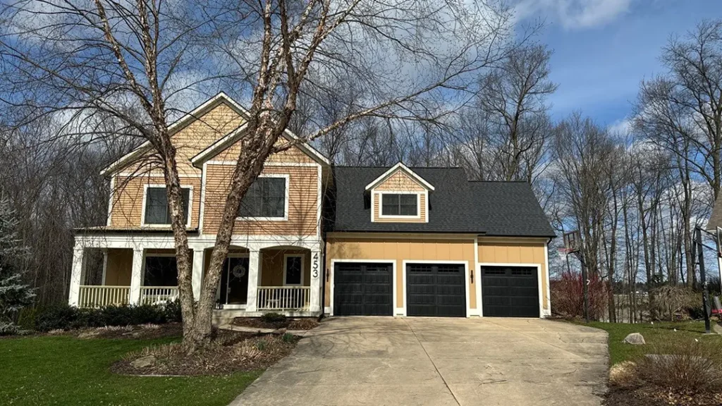 A two-story house with a front porch, three-car garage, and a tree in the yard. The exterior features a combination of brown siding and beige accents. Expertly finished by a trusted roofer, the sturdy roof complements the design perfectly as the driveway extends from the garage.