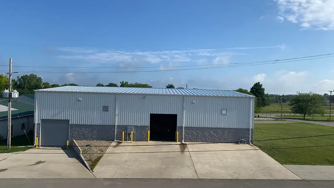 A large, rectangular industrial building with a metal roof and siding, featuring one large garage door and one smaller door. The structure is situated on a paved lot surrounded by grass and trees, showcasing the quality craftsmanship of a top-notch roofing company.