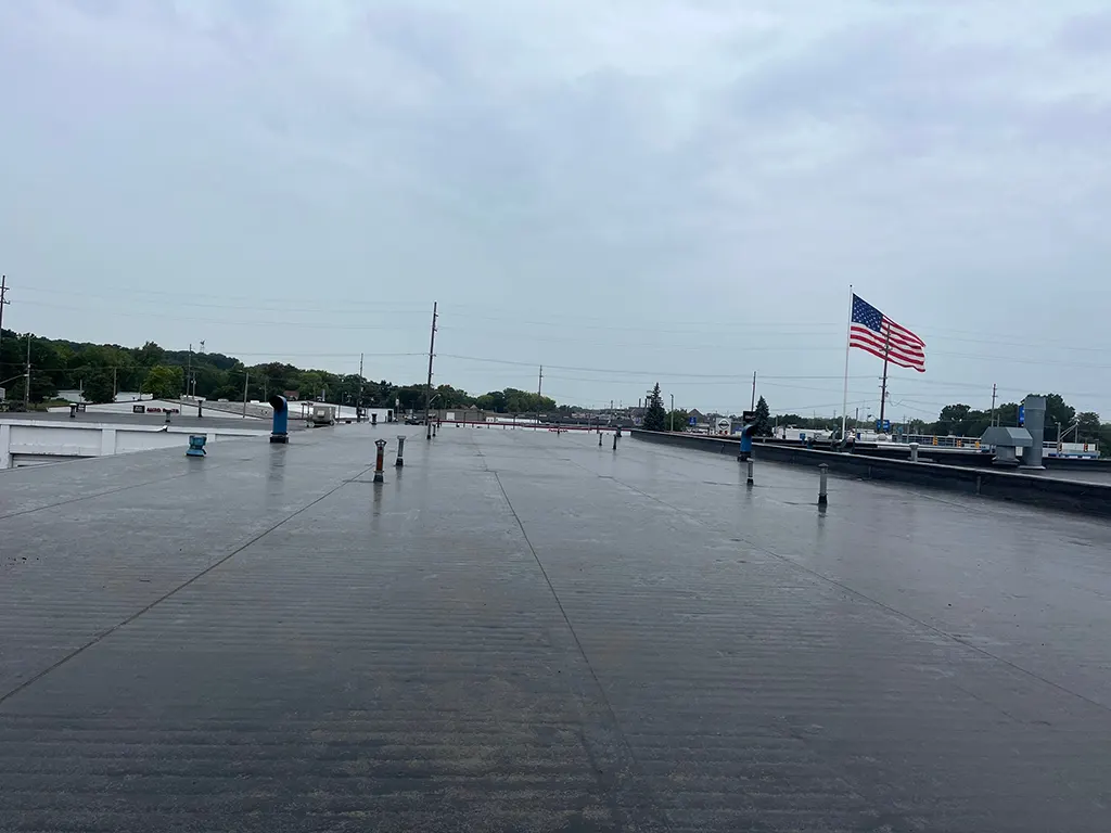 A flat rooftop with various vents and fixtures on a cloudy day. An American flag is visible in the background, flying on a tall pole, showcasing the skilled work of a diligent roofer.