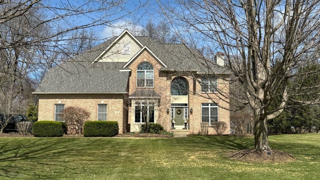 A two-story brick house with large windows and a gabled roof, expertly maintained by a premier roofing company, sits in a yard with leafless trees and a well-kept lawn on a sunny day.