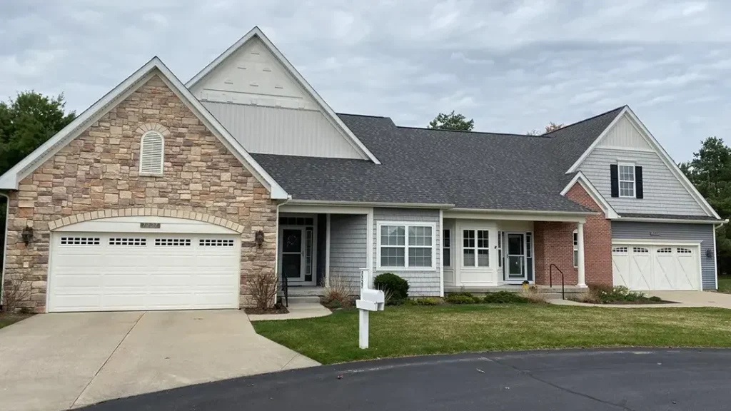 A single-story house with a mix of stone and siding exterior, attached two-car garage, and well-maintained front yard with a mailbox. The driveway leads to a paved street. Recently inspected by a reputable roofing company, the roof adds an extra layer of charm and durability.