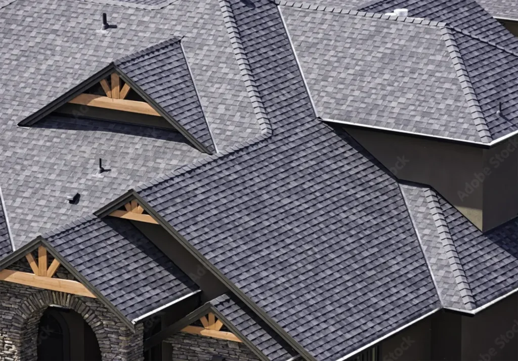 Aerial view of a grey shingled roof on a modern house with multiple gables and dormer windows. The expertly crafted roofing features wooden accents and a stone facade, showcasing the skill of a professional roofing contractor.