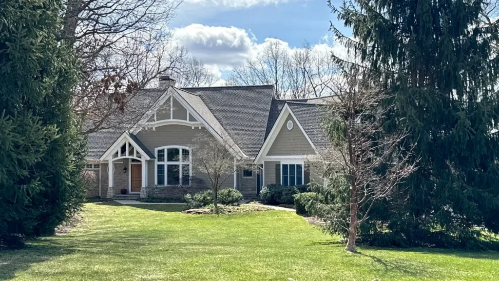 A gray house with white trim and a gabled roof, expertly crafted by top roofing services, is surrounded by trees and a well-maintained lawn under a partly cloudy sky.