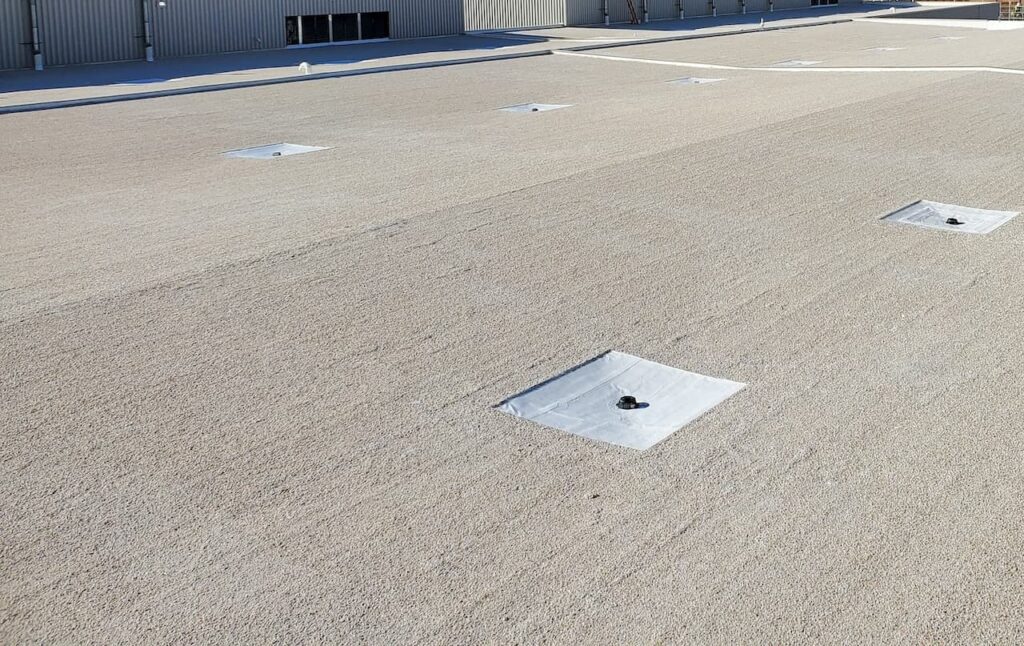 Image shows a large, flat, gravel-covered surface with several square patches of white material, each featuring a black circular object at the center. In the background, metal building structures suggest ongoing work by a roofing company.