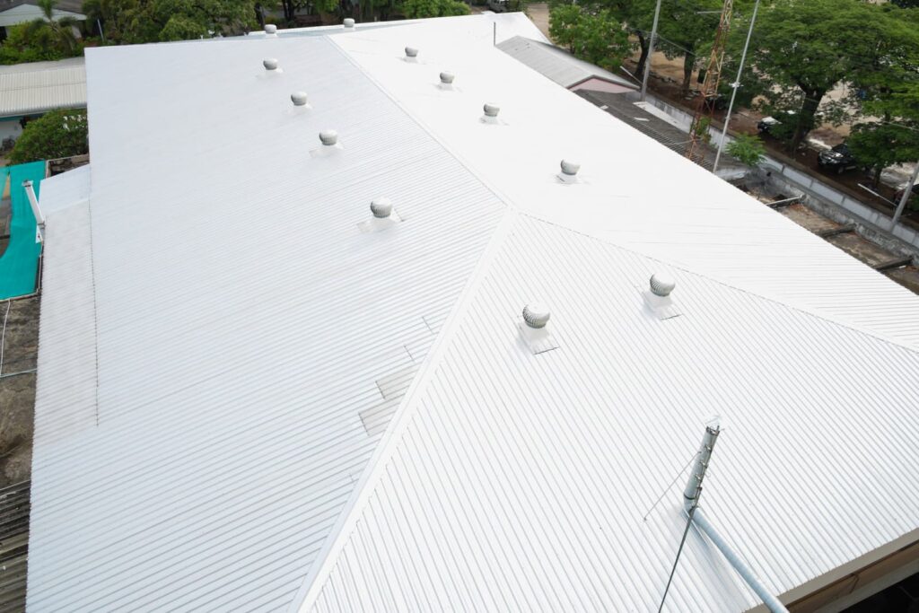 A large white metal roof with several evenly spaced vents, viewed from an elevated angle, showcases the expert craftsmanship of professional roofing services. Trees and neighboring buildings are visible in the background.