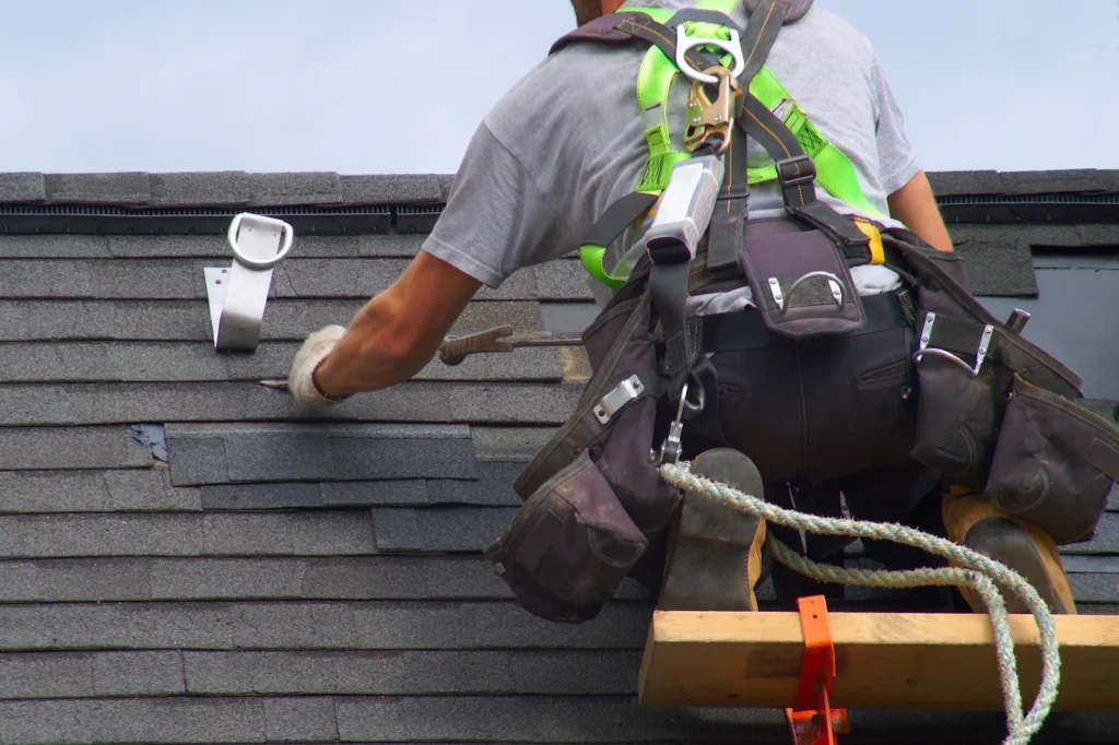 A roofing contractor wearing safety gear repairs shingles on a roof. Various tools are attached to the roofer's belt, and a ladder provides access to the roof.