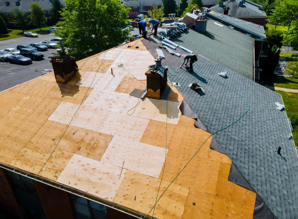 Workers from a roofing company are installing a new roof on a residential building. One half of the roof is covered with new shingles, and the other half has exposed plywood.