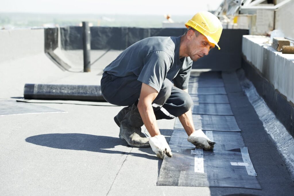 A roofer in a yellow hard hat and gloves installs roofing material on a flat rooftop, showcasing the expertise of their roofing company.
