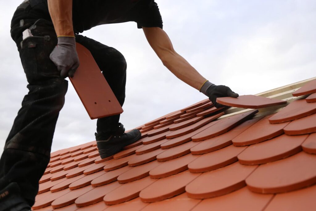 A roofer wearing gloves and holding tiles is installing red roof shingles on a sloped roof, showcasing the expertise of a reputable roofing company.
