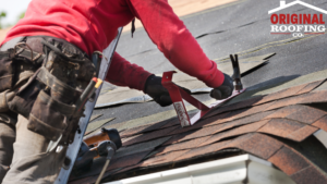 A roofer in a red sweatshirt installs shingles on a roof using a tool. Various equipment is attached to their tool belt. The logo for Original Roofing Co., known for exceptional roofing services, is visible in the top right corner.