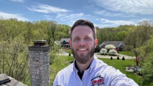 A man in a checkered shirt with "Original Roofing" logo stands outdoors with houses, trees, and a clear blue sky in the background.