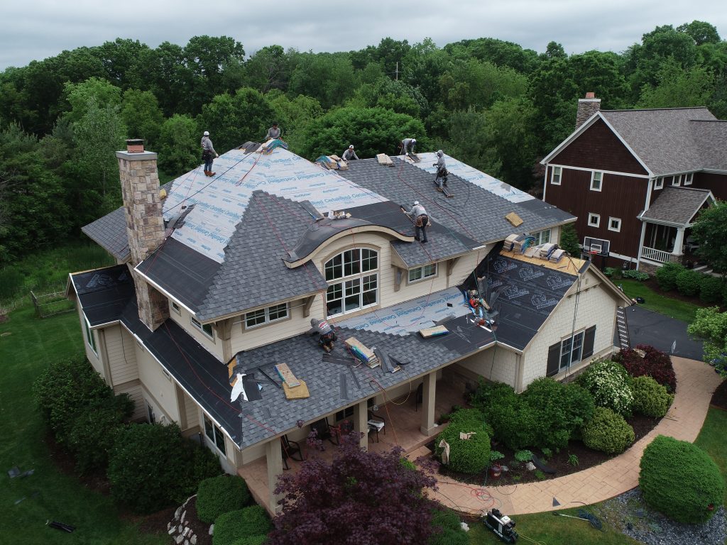 Several workers are installing new shingles on the roof of a large, two-story house surrounded by trees and landscaping. Roofing materials and tools are visible on the rooftop.